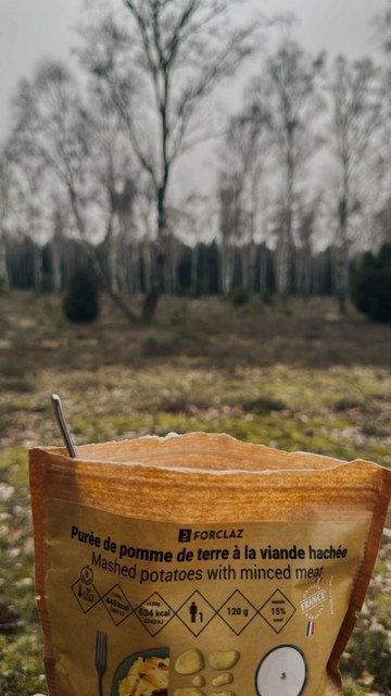 Eine Packung Kartoffelpüree mit Hackfleisch im Vordergrund mit einem unscharfen Hintergrund aus blattlosen Bäumen in einem Wald.