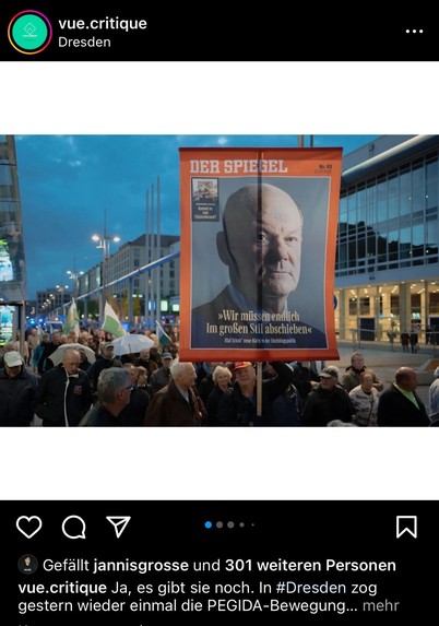 Eine Demonstration mit Menschen, die ein großes Schild in Form der Titelseite des Magazins "Der Spiegel" mit dem Gesicht von Olaf Scholz und dem Zitat „Wir müssen endlich im großen Stil abschieben“, in einer städtischen Umgebung in der Abenddämmerung.