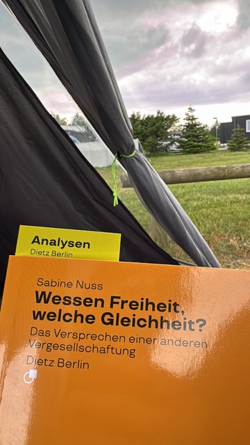 Eine Nahaufnahme eines Buches von Sabine Nuss mit dem Titel "Wessen Freiheit, welche Gleichheit?" in einem Zelt mit einem Hintergrund im Freien aus Gras, Bäumen und einem bewölkten Himmel.