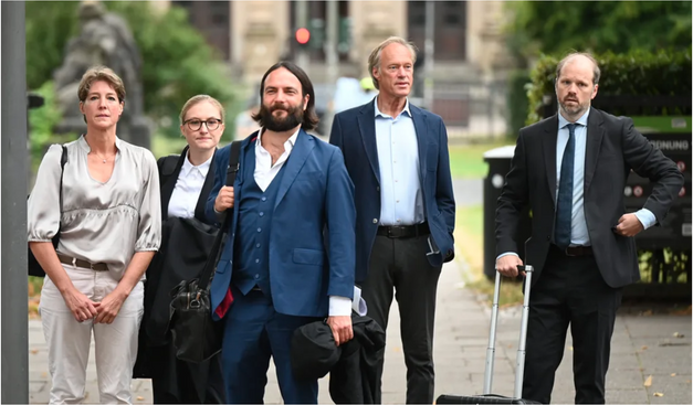 Ein Pressefoto von Christina Block, Gerhard Delling und deren Anwält*innen. Im Vordergrund steht der Anwalt Ingo Bott und lächelt verschmitzt in die Kamera