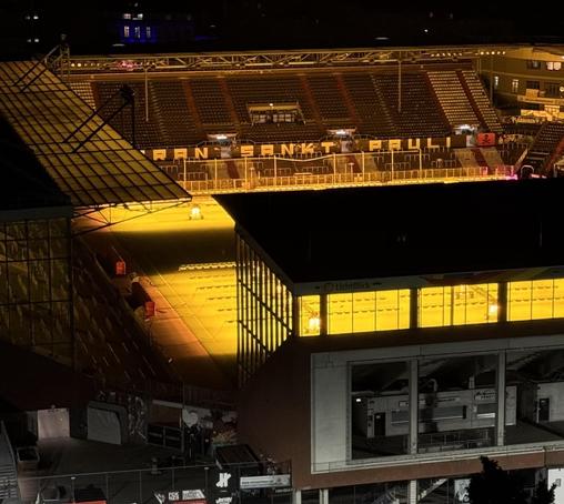Ein nächtlicher Blick auf das teilweise beleuchtetes Millerntorstadium mit Sitzgelegenheiten und einem von Natriumdampflampen hell gelb beleuchteten Spielfeld. Der Name "FC St. Pauli" ist auf den Sitzplätzen zu sehen.