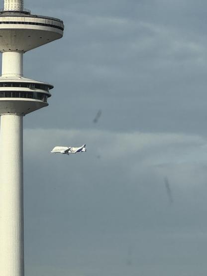 Ein Foto, das in Vordergrund den Hamburger Fernsehturm zeigt. Rechts davon fliegt ein Airbus Beluga (Transportflugzeug). Durch den Zoom sieht es ein bisschen so aus als würde das Flugzeug auf den Turm zufliegen.