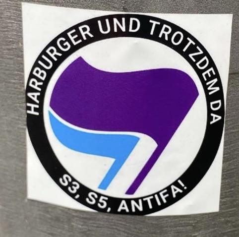 Ein runder Sticker den zwei Flaggen der antifaschistischen Aktion (eine lila, eine hellblau“ und drum herum der Spruch „Harburger und trotzdem da - S3 S5 Antifa“