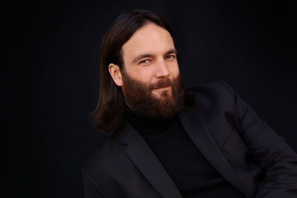 Ein Bild von Anwalt Ingo Bott. Er sitzt in schwarzem Anzug vor einem schwarzen Hintergrund. Er trägt einen Vollbart und Haare, die bis zu den schultern reichen und schaut dazu geradezu lasziv in die Kamera