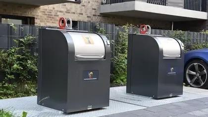 Zwei Abfallbehälter mit gewölbten Deckeln stehen nebeneinander auf einem gepflasterten Untergrund, vor dem Hintergrund von Grünflächen und einem Gebäude.