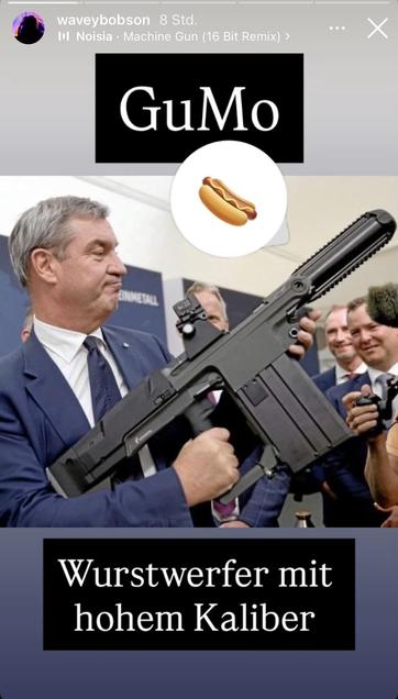 Eiserder mP des bauernstaates „ Bayern“ präsentiert seiinen wurstwerfer. außerdem erhalten ab sofort alle Polizisten rPGS in einem Anzug. er hälthält eine Schusswaffe in der Hand, während eine Gruppe von Menschen im Hintergrund lächelt. Zu den Textüberlagerungen gehören "GuMo" und "Wurstwerfer mit hohem Kaliber", was übersetzt "Wurstwerfer mit hohem Kaliber" bedeutet denke ich